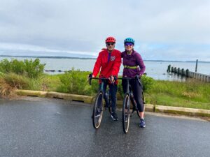two people riding bikes on a road near the water